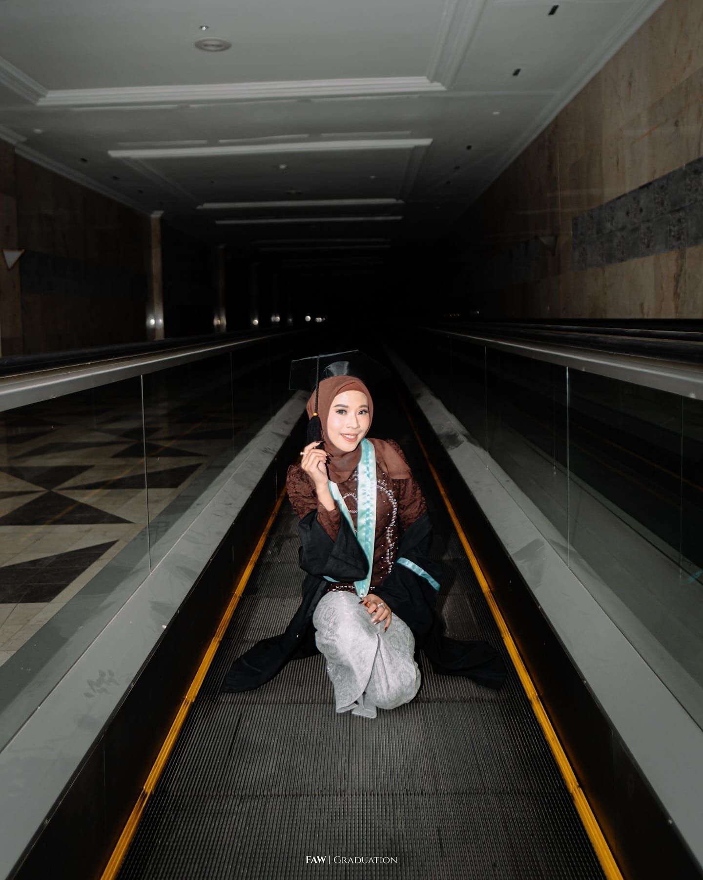 Wisudawati bertoga duduk di travelator saat sesi malam, garis kuning eskalator menyala di sekitarnya