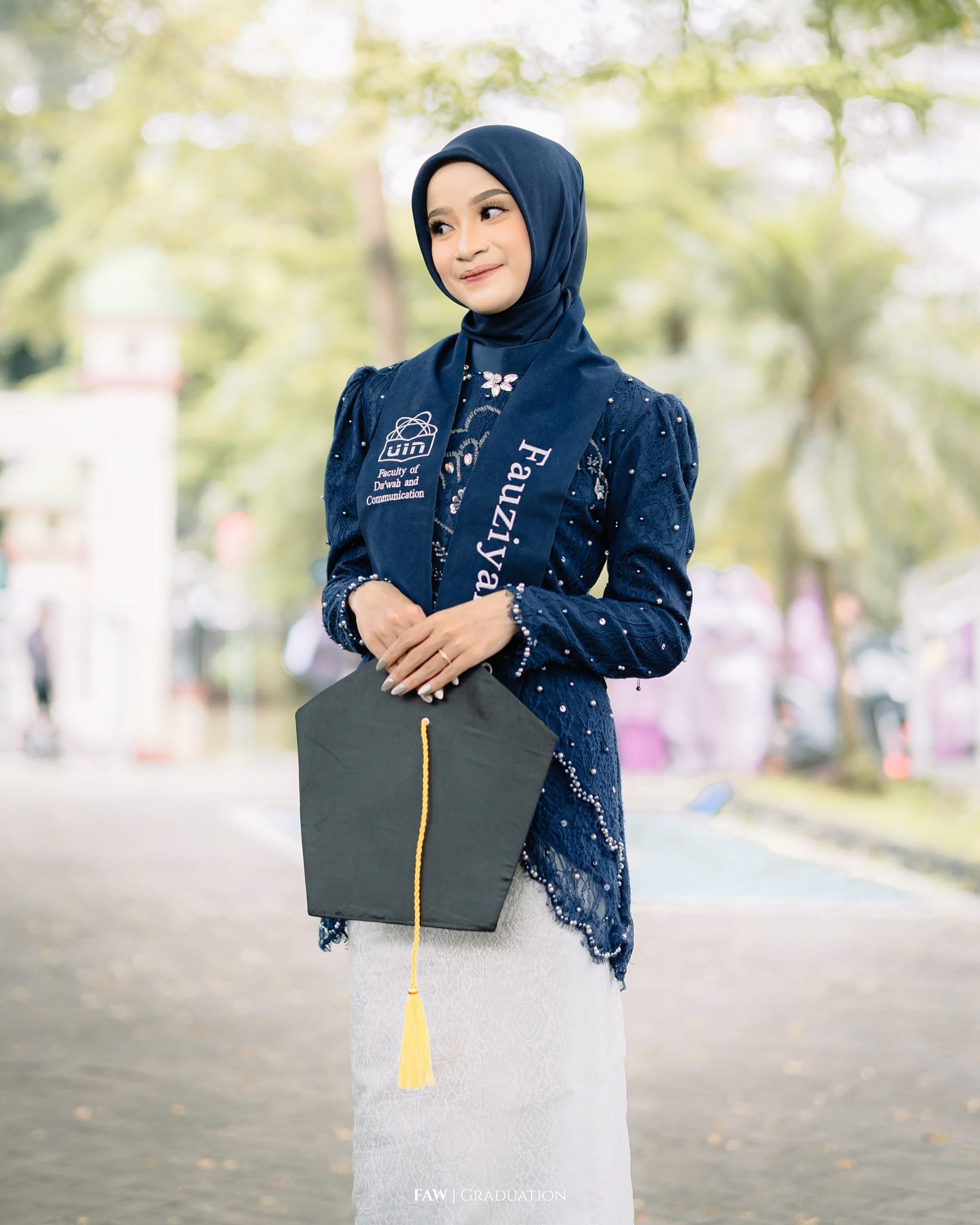 Wisudawati berkebaya biru dongker memegang toga, selempang Fakultas Dakwah dan Komunikasi UIN Jakarta