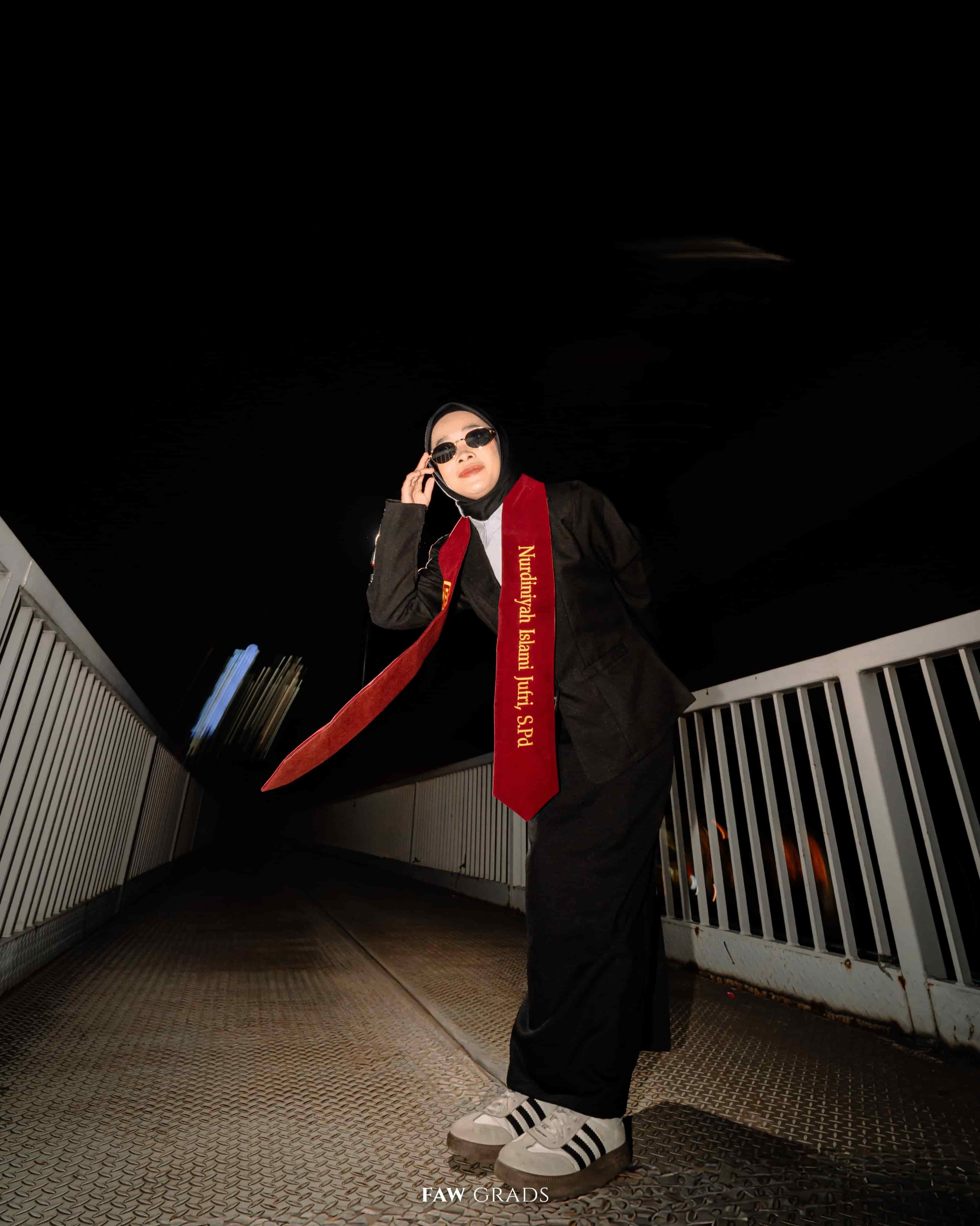 Wisuda malam di jembatan kota: wisudawati berhijab bertoga hitam, selempang merah, kacamata hitam, dan sepatu kets, bergaya urban