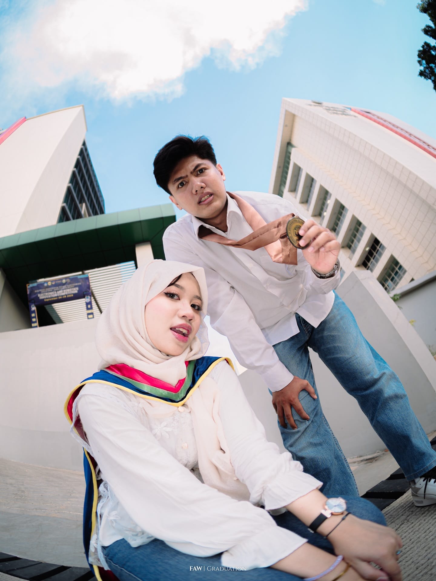 Dua sahabat berpose di depan gedung kampus dengan medali dan selempang, dipotret dengan lensa fisheye